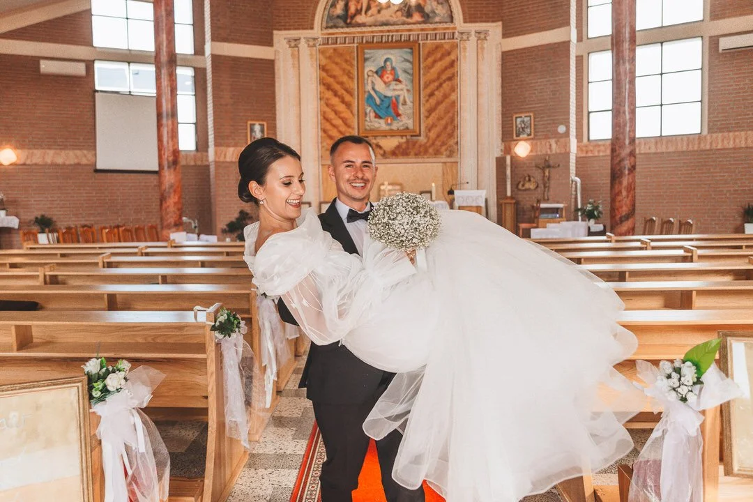 A bride and groom inside a church, with the groom carrying the bride in his arms. The bride is holding a bouquet of white flowers, and both are smiling. The church has wooden pews, brick walls, and religious artwork in the background.