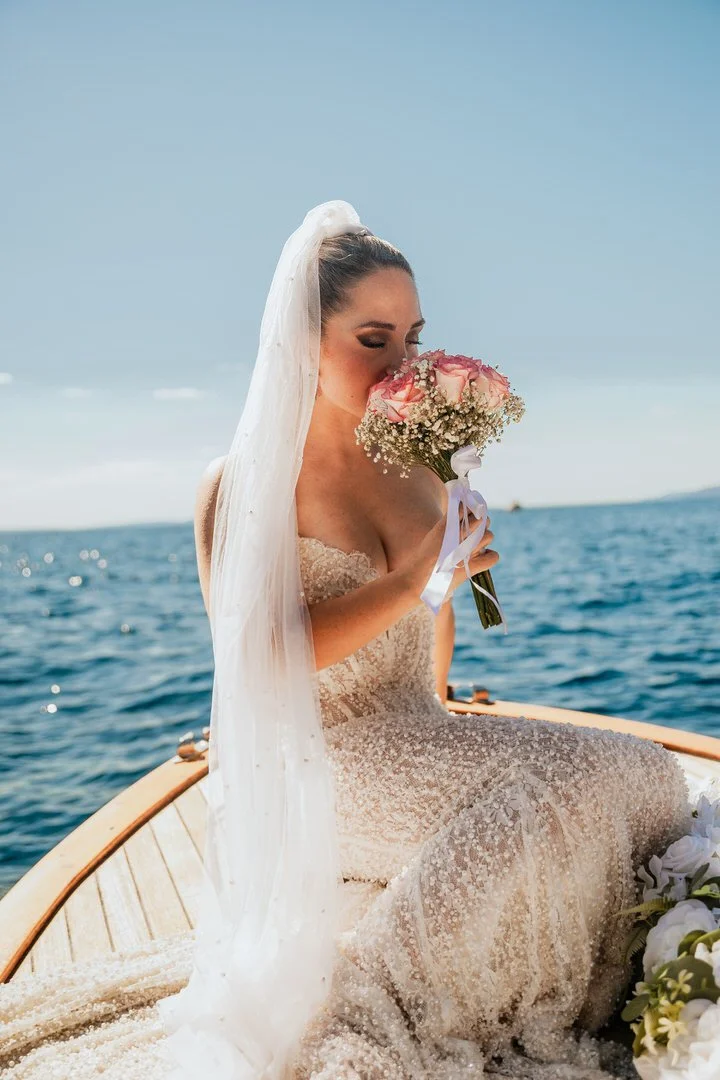 Bride in a wedding dress and veil sitting on a boat in Kvarner Bay near Opatija, holding a bouquet of pink roses and baby's breath during a wedding photoshoot.