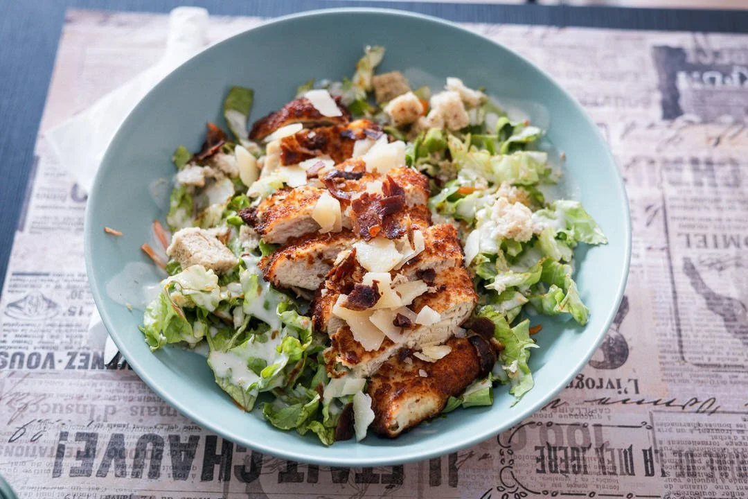 Bowl of Caesar salad with grilled chicken, chopped lettuce, shredded cheese, and croutons on a table with newspaper print.