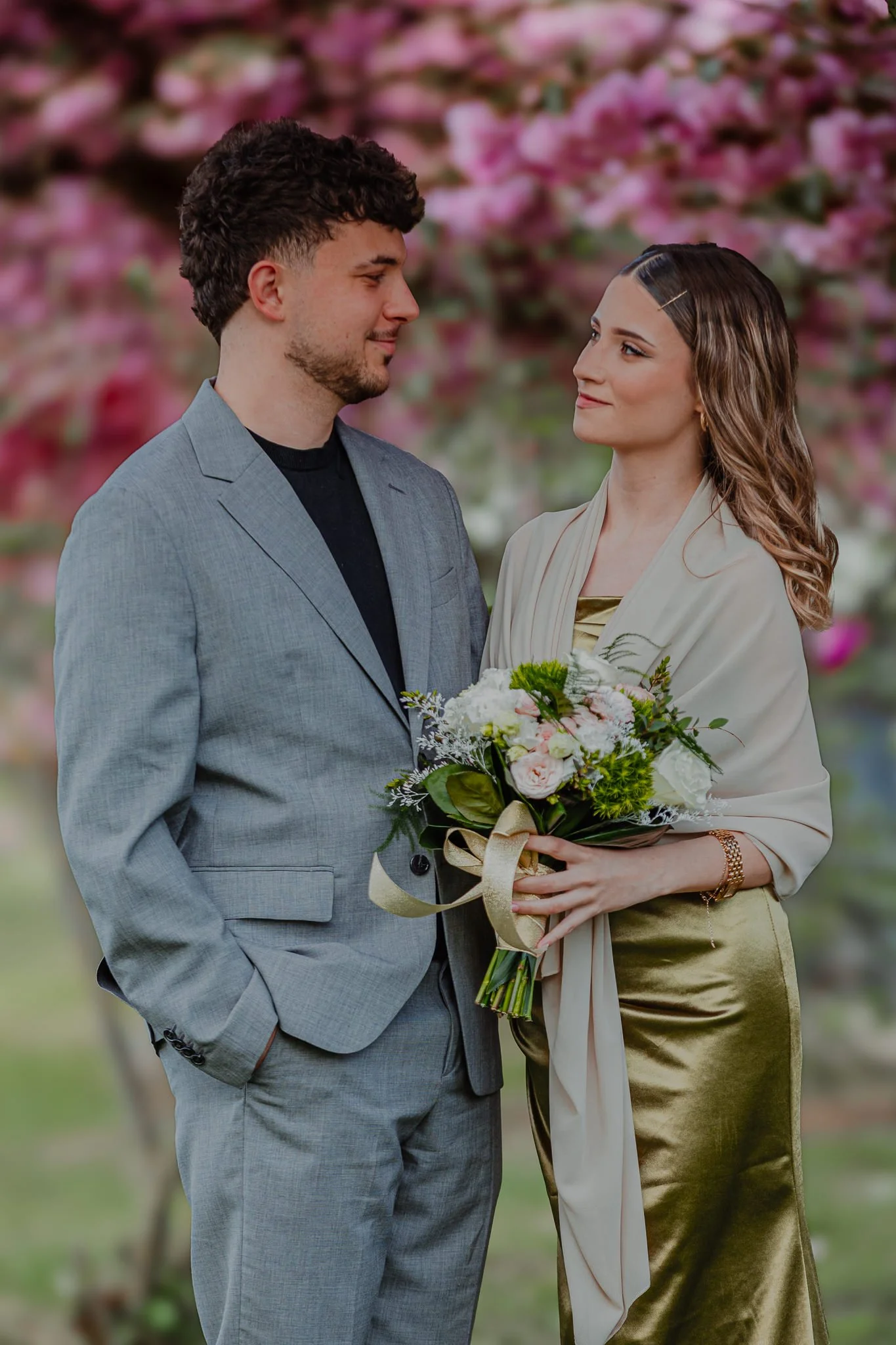 A man and woman are standing close together outdoors, facing each other, with pink flowering trees in the background. The woman is holding a bouquet of flowers.