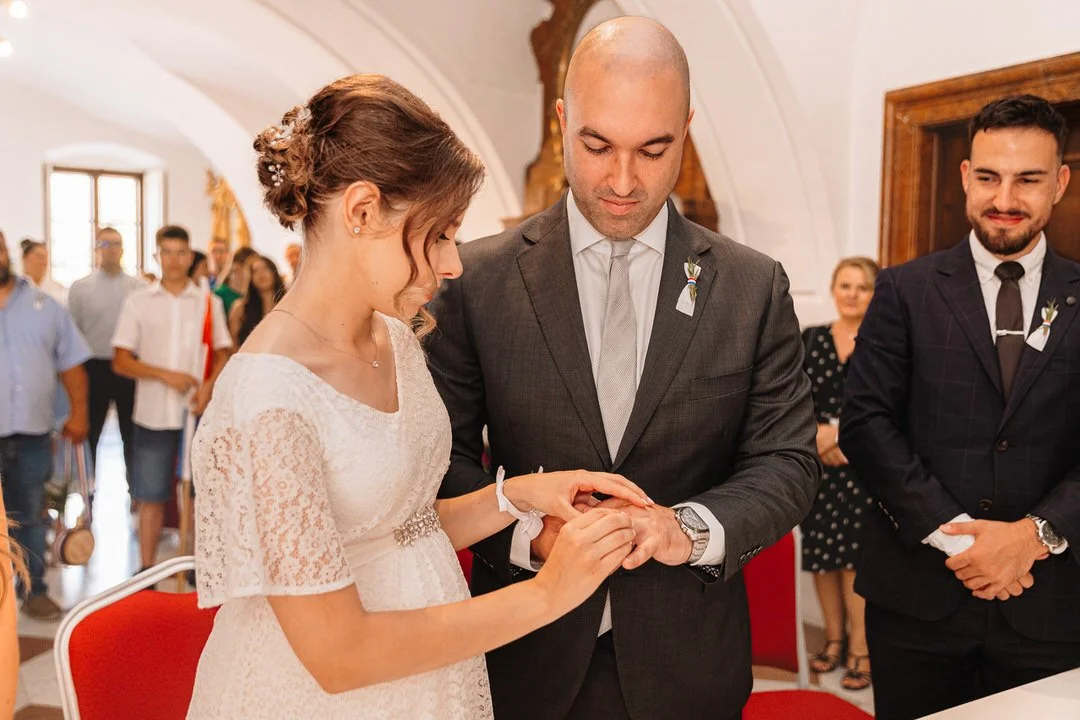 A couple is participating in a wedding ceremony in Vukovar, with the bride placing a ring on the groom's finger in front of guests.