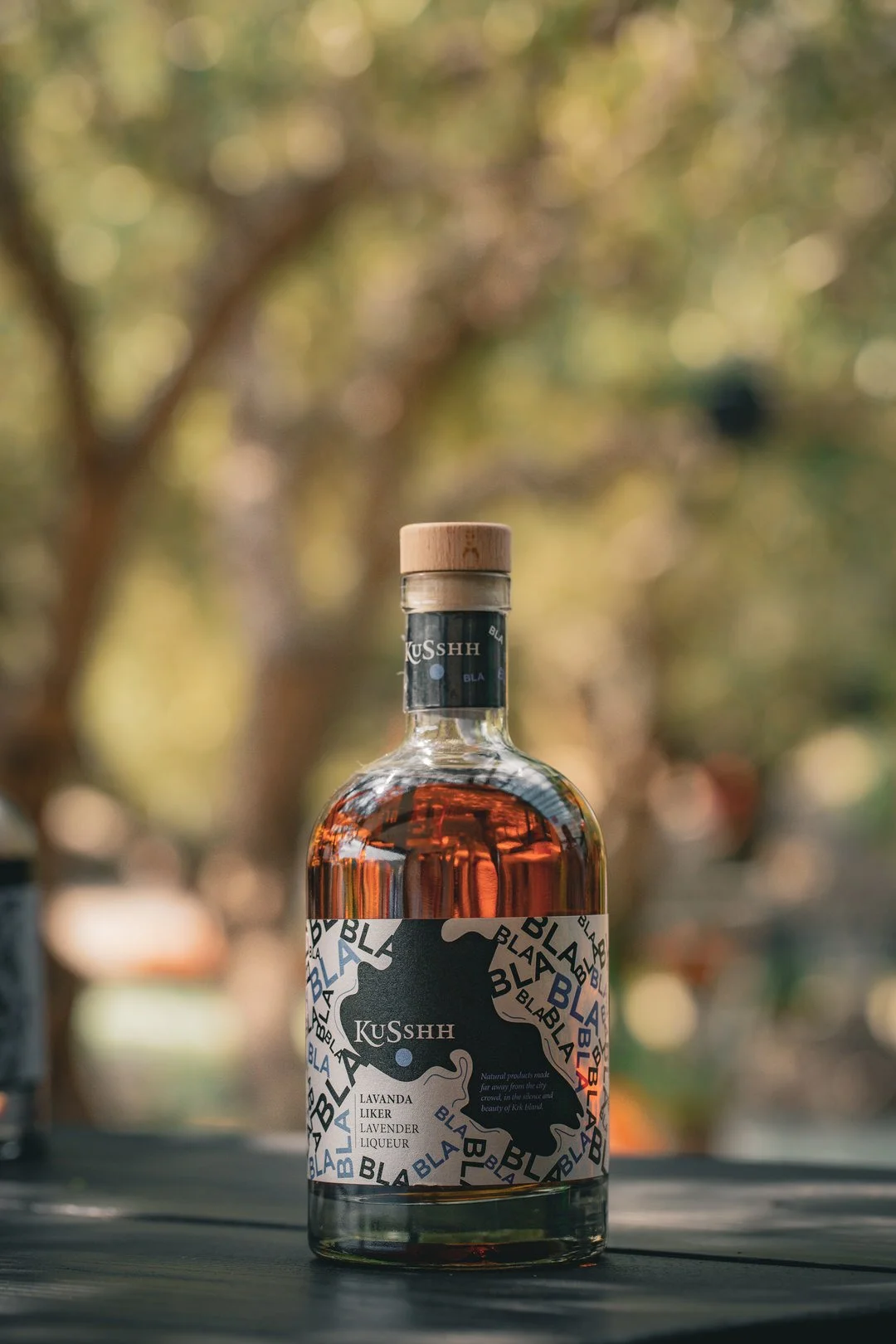 A bottle of lavender liqueur with a black matching label and a light wooden cap, placed outdoors on a dark surface with blurred trees and sunlight in the background.