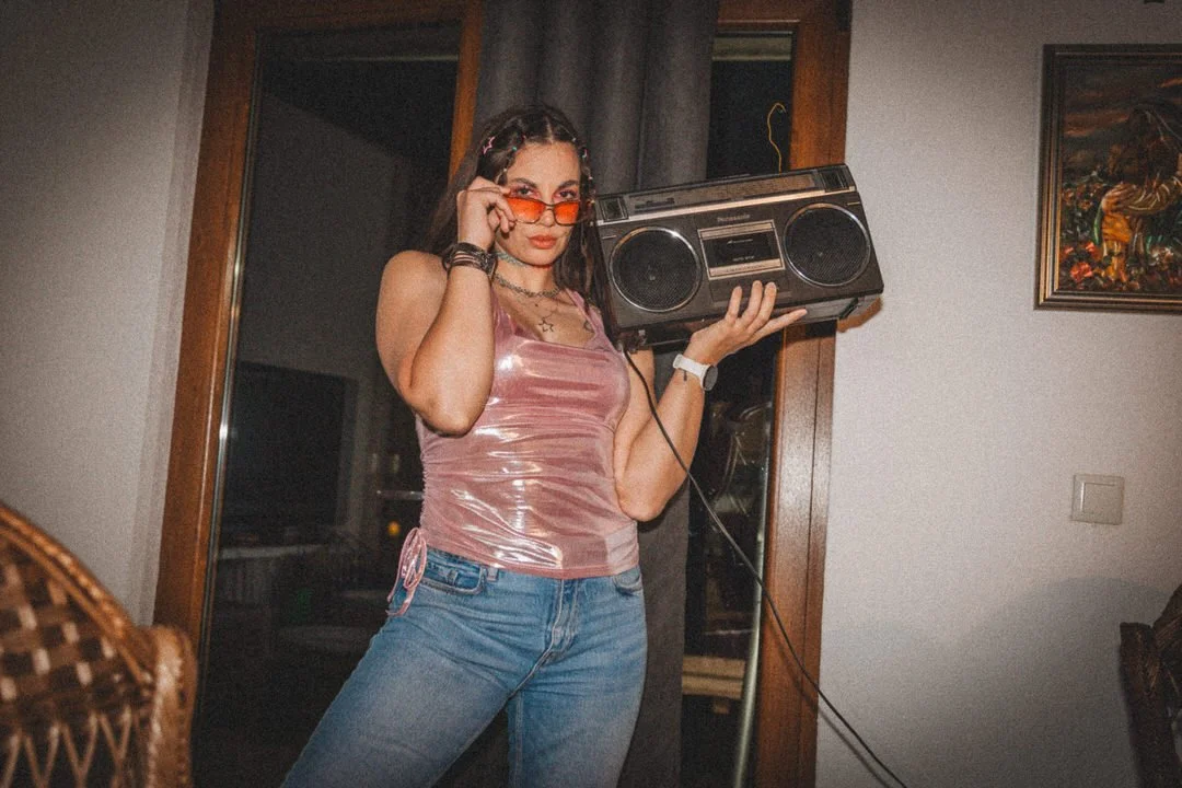 A woman wearing a shiny pink tank top, jeans, and red-tinted sunglasses holding a vintage boombox and posing indoors.