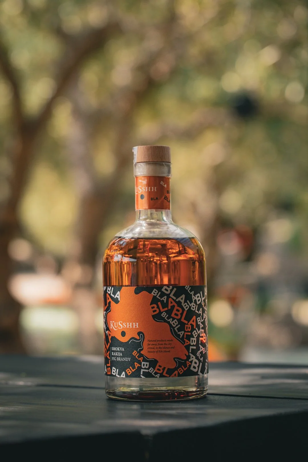 A bottle of KuSshh Fig Brandy with a black and orange label, placed outdoors on a wooden surface with a blurred background of trees and sunlight at Ulika Grill Restaurant, Vrbnik, Krk Island, Croatia.