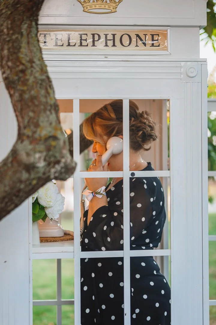 A woman is talking on a vintage white telephone inside a white phone booth, partially obscured by a tree in the foreground. She is wearing a black dress with white polka dots and has short, wavy hair. A small bouquet of white flowers is visible near 