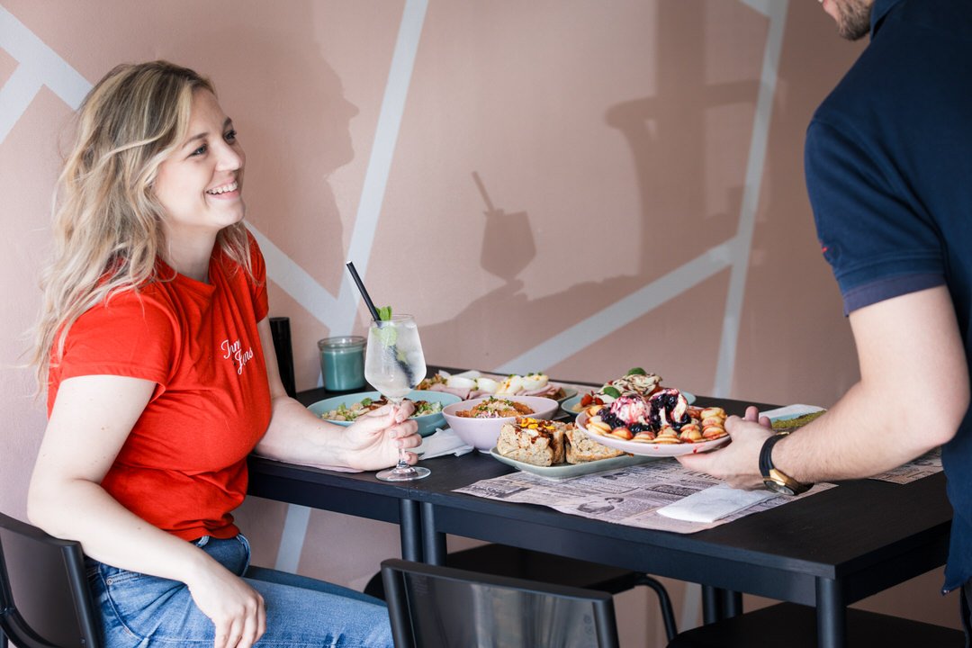 A woman in a red shirt sitting at a table, smiling as a man serves her a plate of desserts and food, with various dishes and a drink on the table.