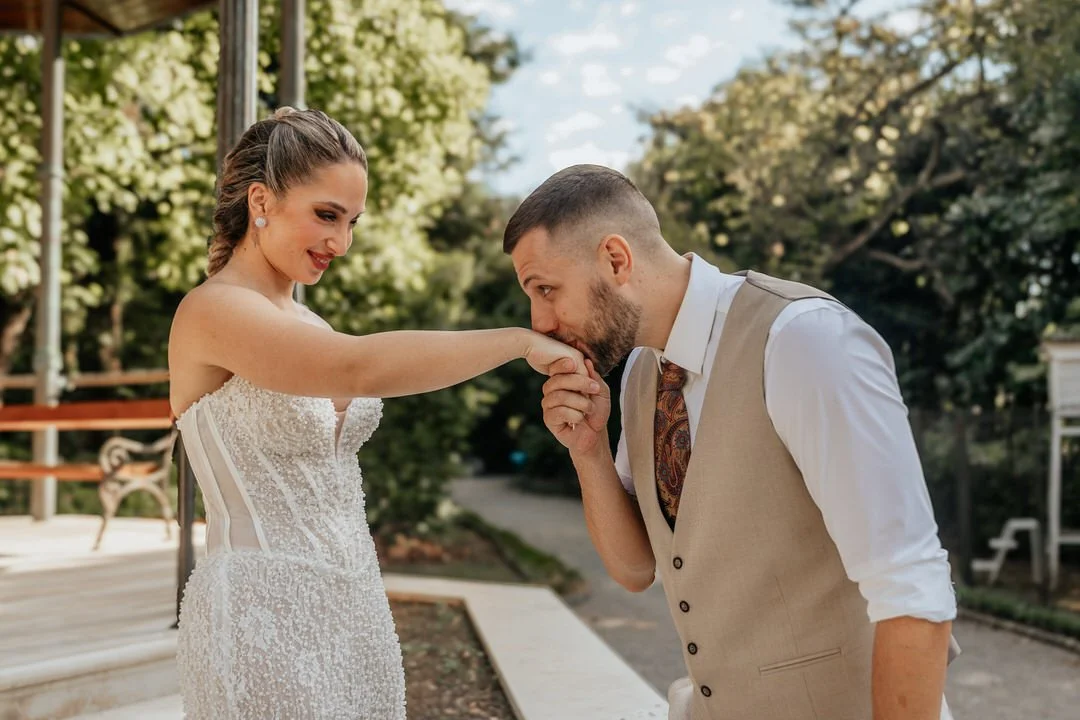 A bride and groom outdoors, with the groom kissing the bride's hand.