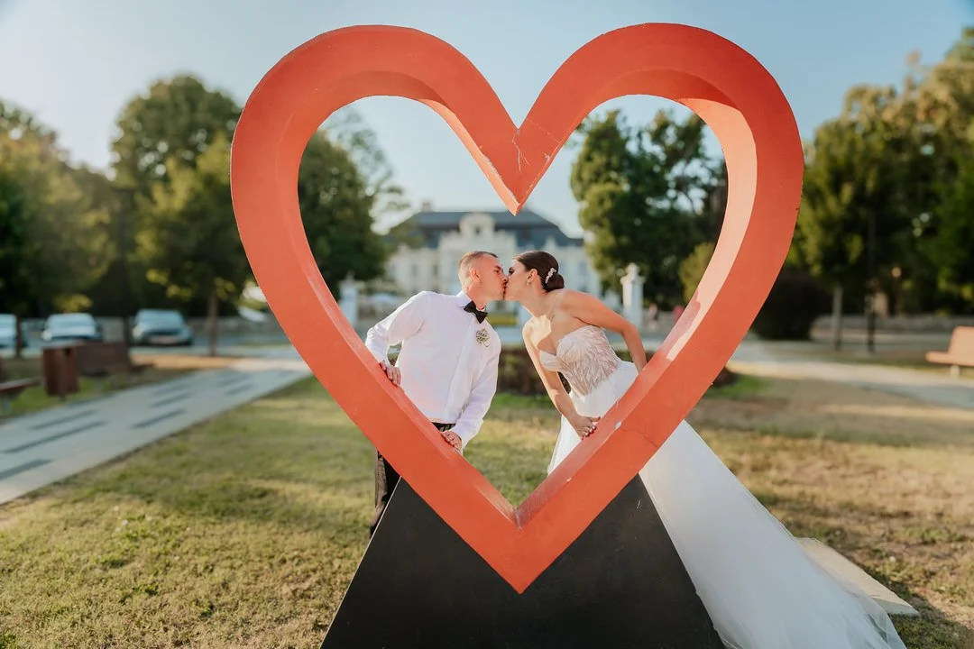 A bride and groom kissing inside a large red heart-shaped frame on a grassy area in Našice during their wedding day.