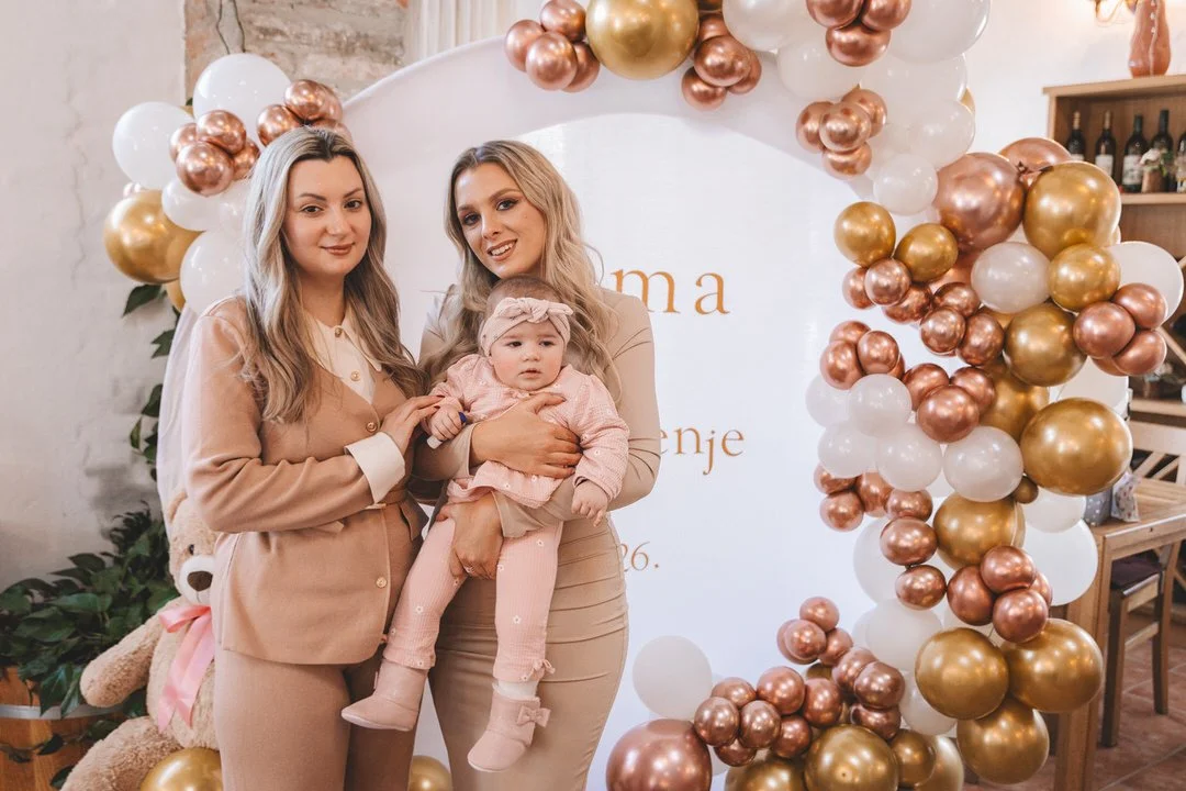 Three females standing in front of a balloon arch at a baptism celebration, smiling and dressed in coordinated outfits.