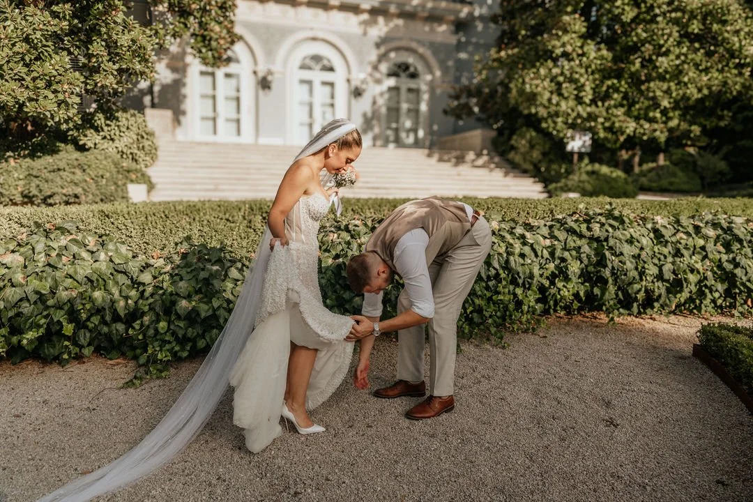 A bride in a wedding dress and veil is adjusting her shoe while a man helps her, near a garden with green bushes and large building in the background.