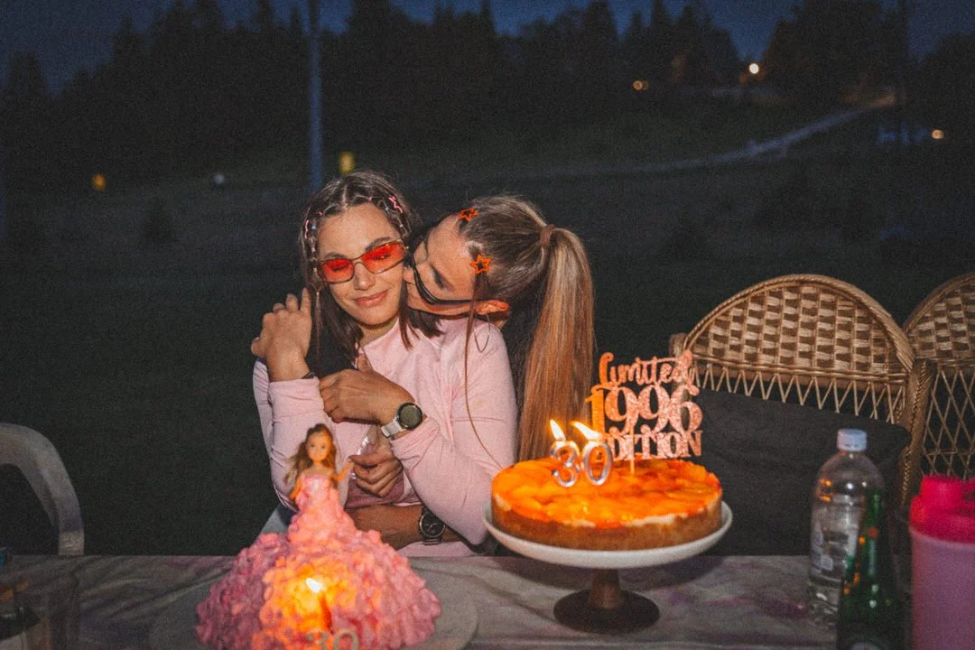 Two young women celebrating a birthday outdoors at night, with a cake that has candles and a topper reading "Cumpleaños 1996" and the number "30". One woman is hugging the other, with a pink dress, and they are sitting at a table with other desserts 