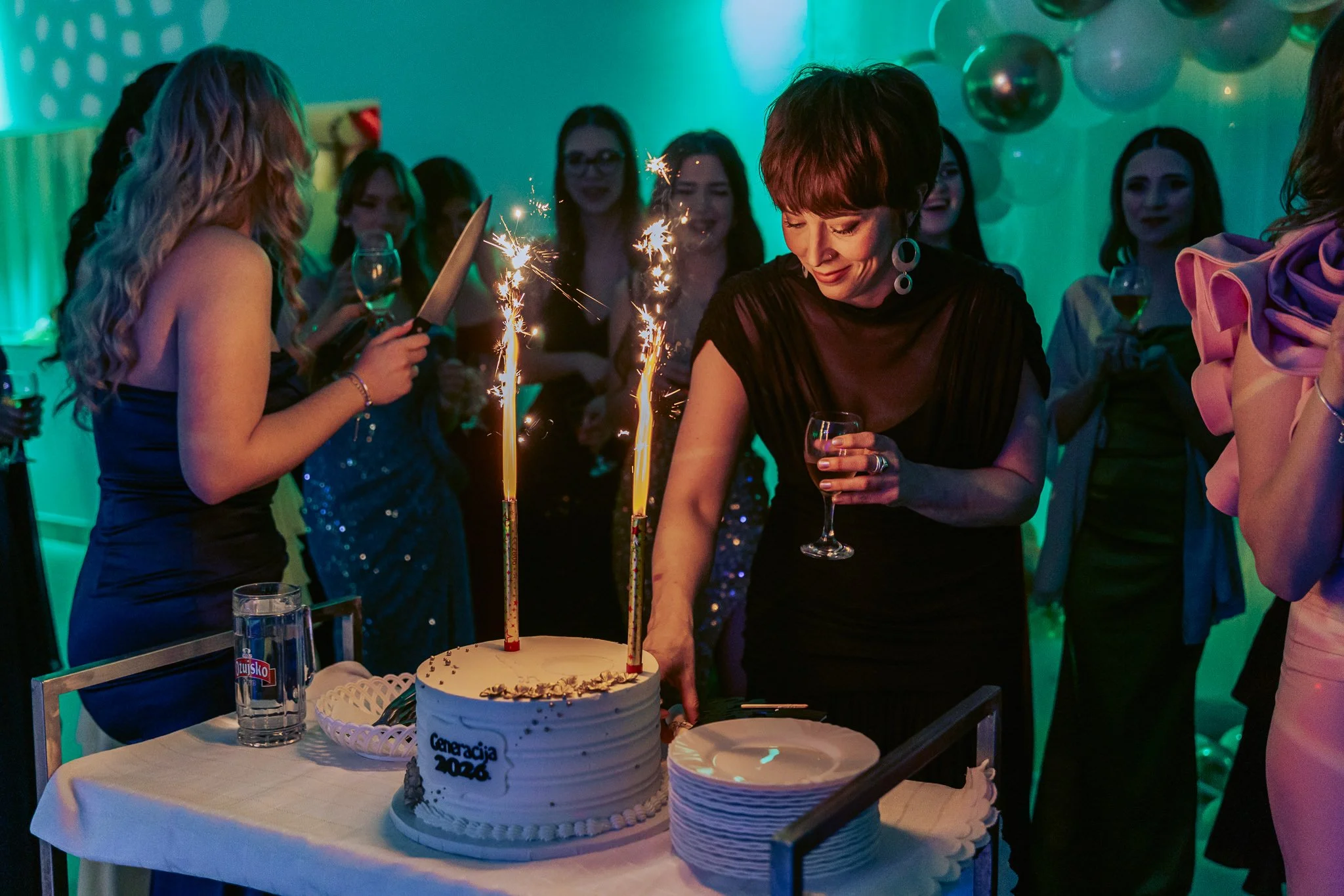 A birthday prom celebration with a cake, sparklers, and several women gathered around, smiling, at a party with green and blue lighting, balloons, and drinks.