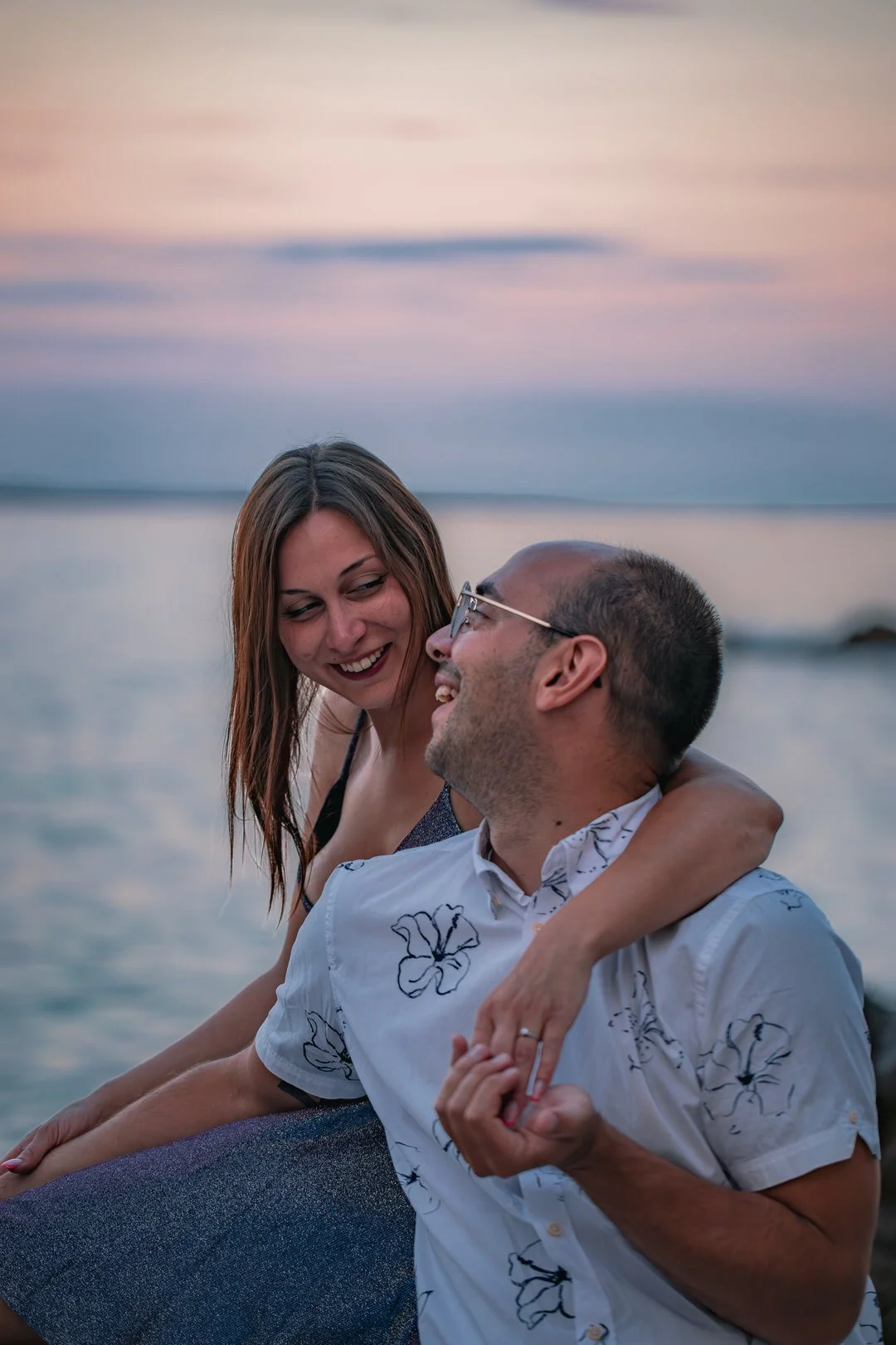 A woman and a man are sitting by the Adriatic Sea during sunset, smiling at each other during an engagement session on Krk Island, Croatia. The woman has her arm around the man's shoulders.