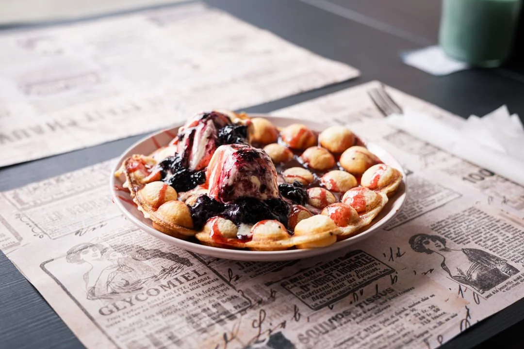 A plate of waffles topped with strawberries and blueberry syrup on a piece of newspaper-style parchment paper.