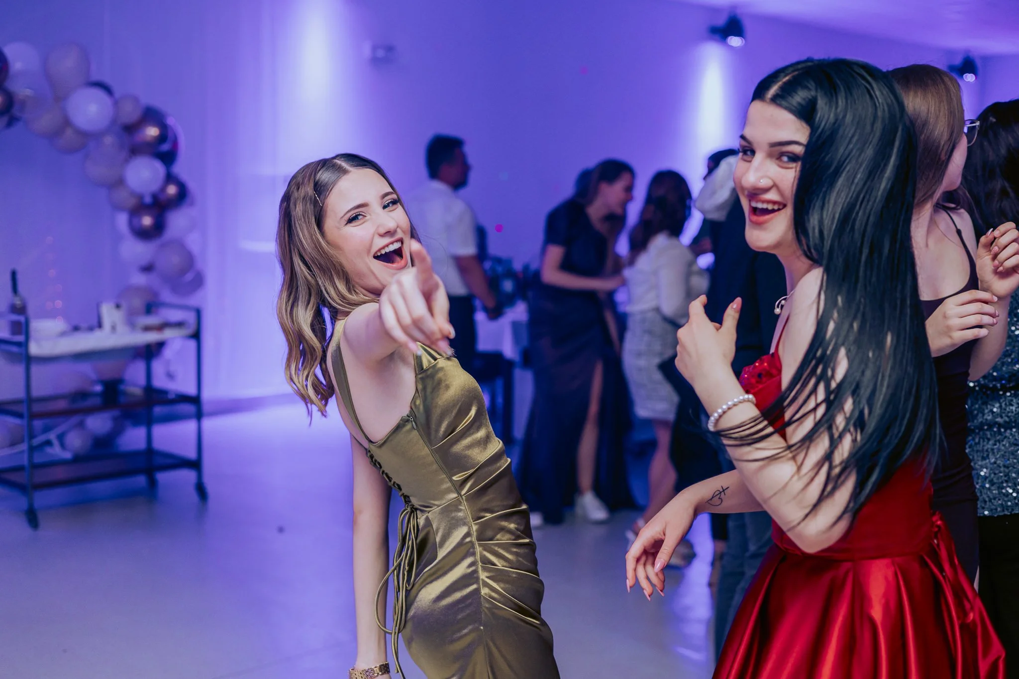 Two women dancing and smiling at a party with purple lighting, balloons, and guests in the background.