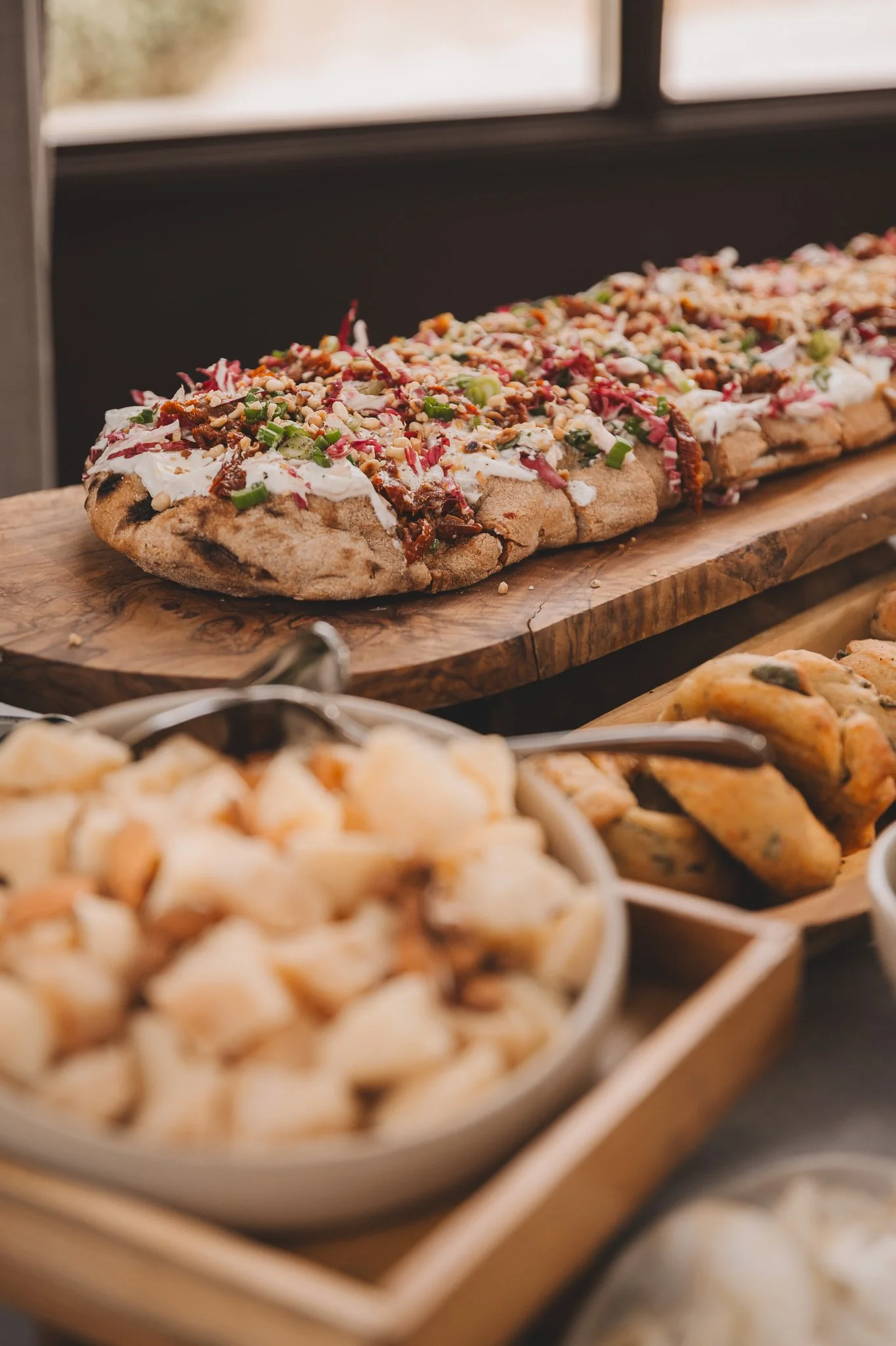 Close-up view of a decorated flatbread topped with green onions, bacon, and chopped nuts on a wooden board, with other baked goods in the foreground and window in the background.