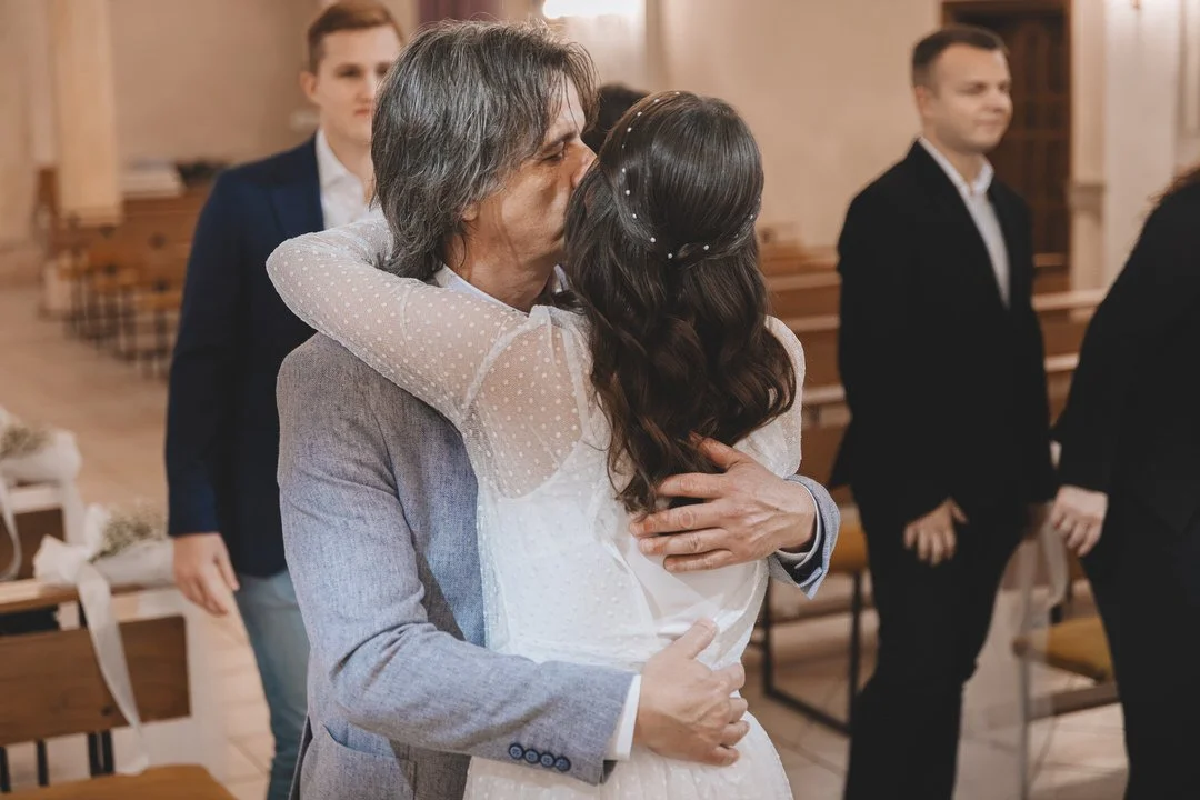 A bride hugging her father during her wedding ceremony, sharing an emotional moment, with other guests in formal attire in the background.