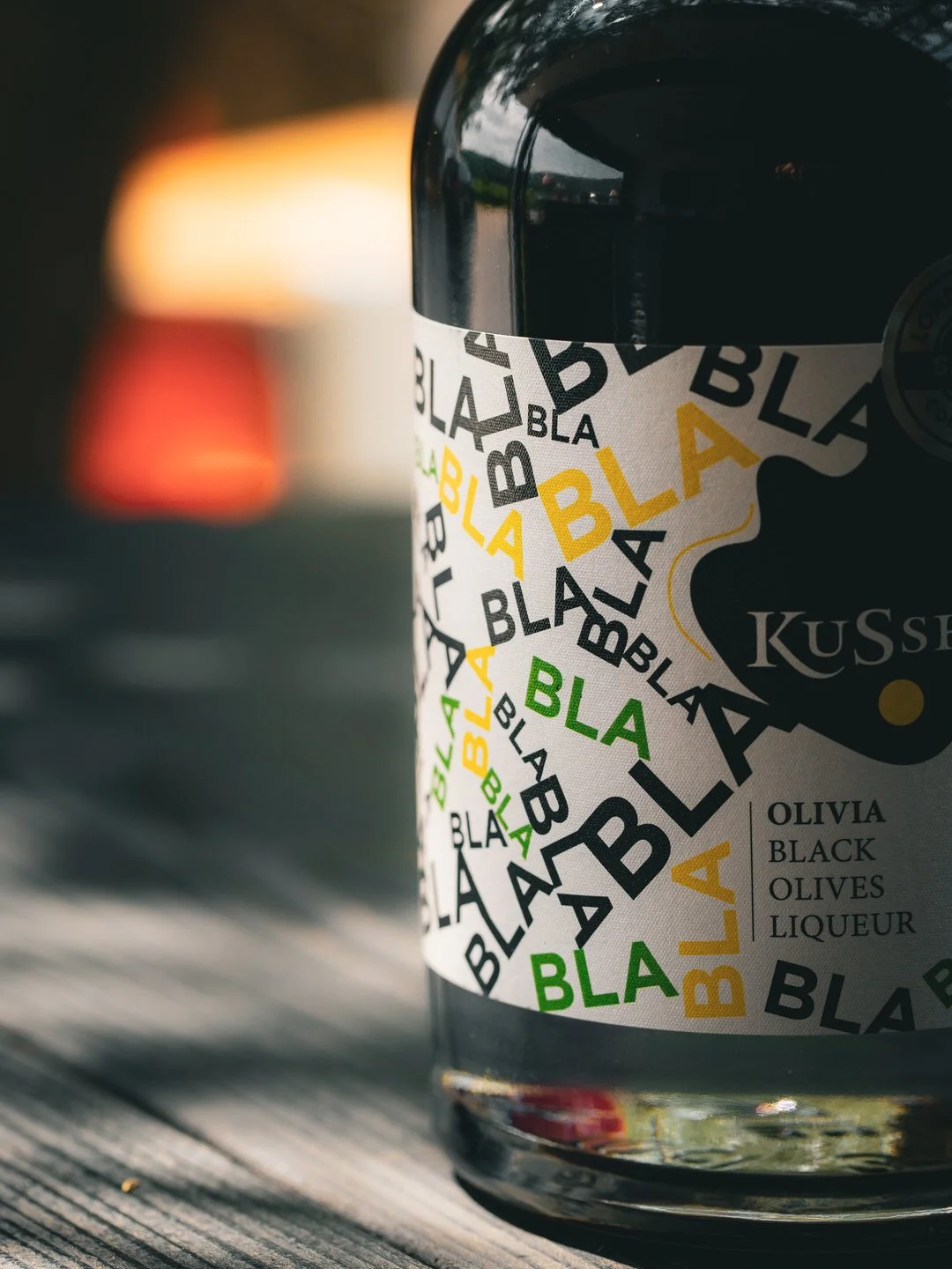 Close-up of a glass jar with a label that reads "Olivia Black Olives Liqueur" in black, green, and yellow text, placed on a wooden surface with blurred background.