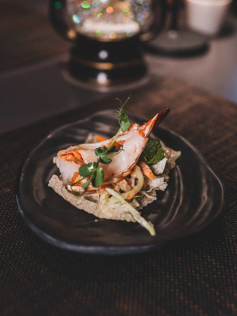A plate of gourmet shrimp cocktail garnished with microgreens, served on a black plate with a textured rim, set on a dark tablecloth in a dimly lit setting.