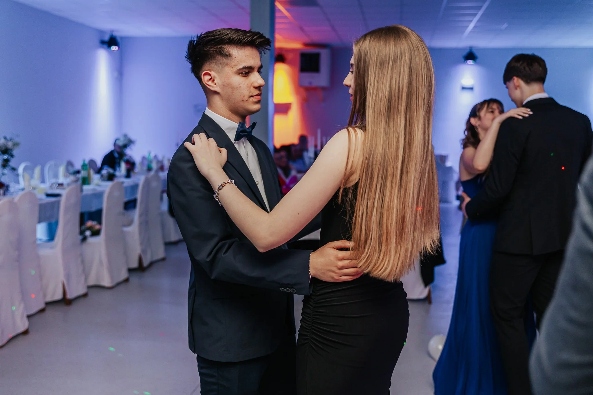 Couple dancing closely at a formal event or wedding reception, with other couples dancing in the background and decorated tables with chairs.
