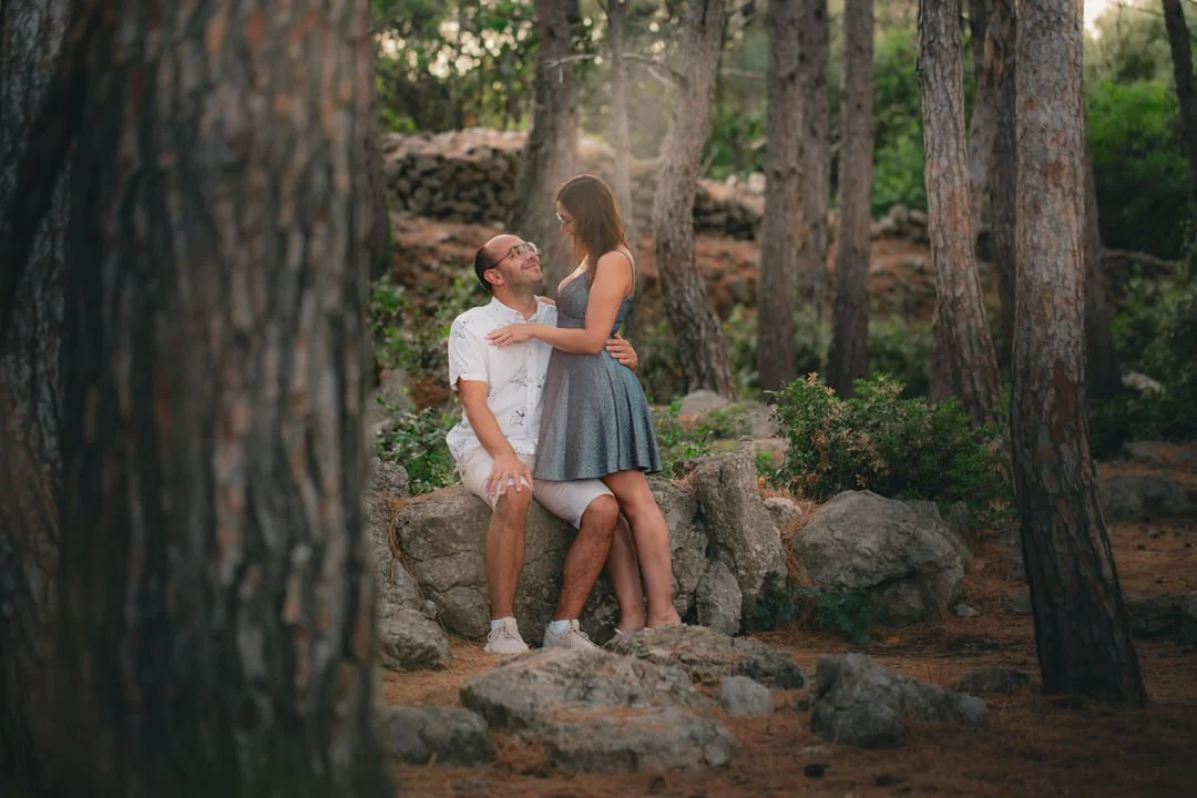 A couple sitting on a large rock in a forest, with trees and greenery in the background, sharing a tender moment at Plav Beach, Krk Island, Croatia.
