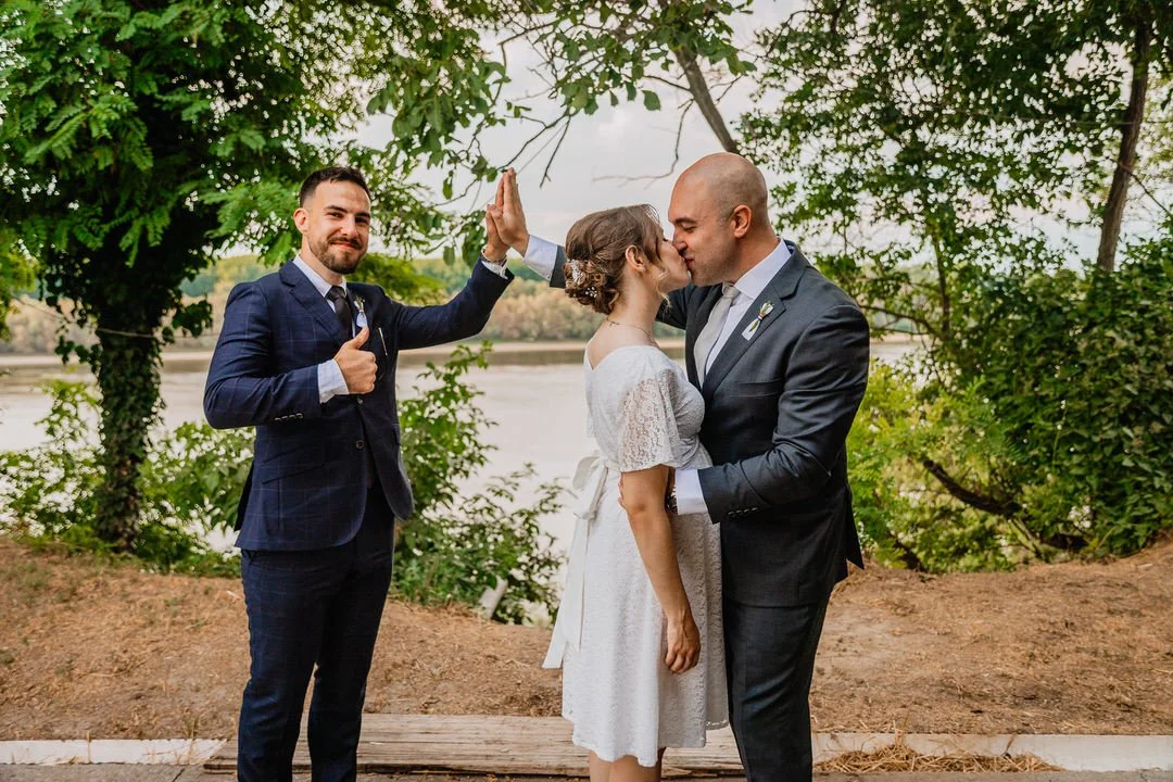 Bride and groom kissing outdoors near a lake, a best man giving a thumbs up and high-five.