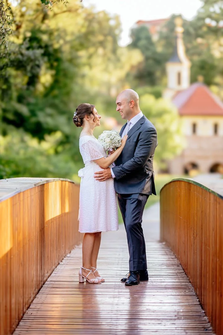 A bride and groom standing on a wooden bridge in park forest Adica, smiling at each other, with the bride holding a bouquet of flowers, outdoors with greenery and a building in the background.