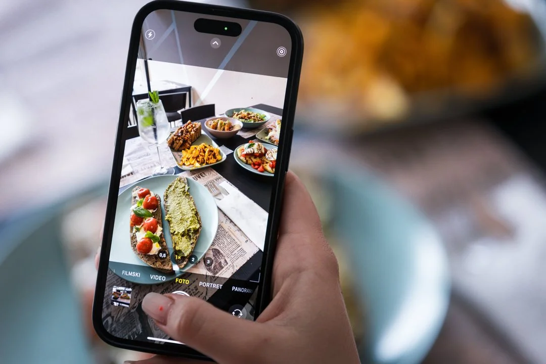 A person holding a smartphone capturing a photo of a table with various foods, including a plate with bread topped with tomatoes and basil, and several bowls of different dishes.