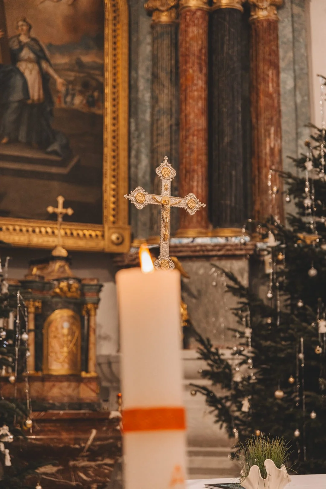 A lit white candle with an orange ring around the bottom and a decorative gold cross in the background during a Croatia baptism in Slavonija, set inside a church with religious artwork and Christmas decorations.