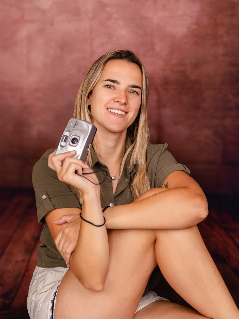 A young woman with blonde hair, smiling and holding a small silver camera, sitting with her arms crossed and legs bent, against a blurred brown background.