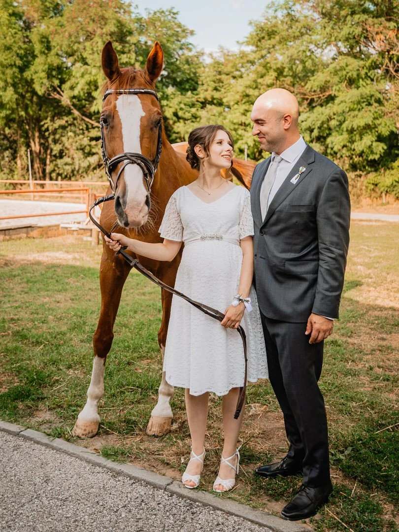 A couple dressed in wedding attire standing outdoors with a brown horse, trees, and a wooden fence in the background in Dunavski Raj venue in Vukovar