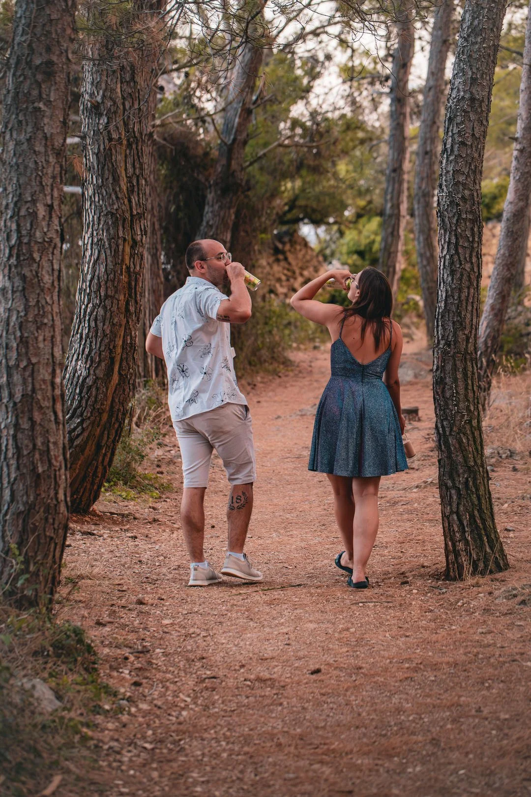 A man and woman walking on a dirt trail in a wooded area, drinking from cans during sunset, during an engagement session.