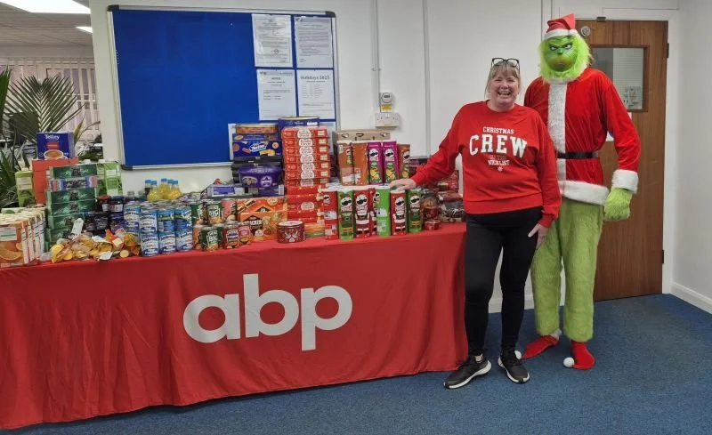 Woman in red Christmas crew sweatshirt standing next to a person in a Grinch costume, both standing behind a table filled with Christmas food items, in a room with white walls and a blue bulletin board.