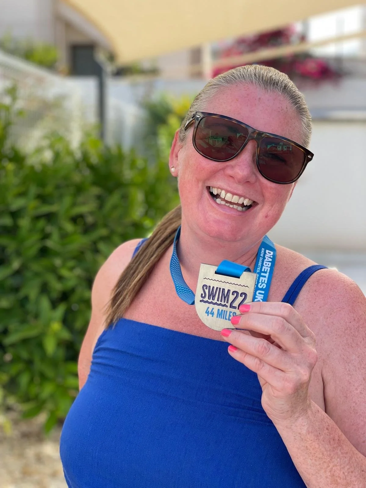 Smiling woman in blue top holding a swim race medal reading 'SWIM 22 44 MILES' with a blue ribbon, outdoors with green foliage and a building in the background.