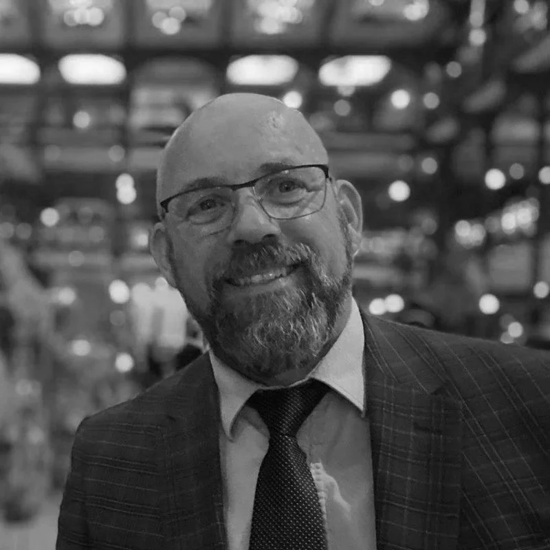 A smiling man with glasses and a beard, wearing a suit and tie, in a setting with blurred lights in the background.