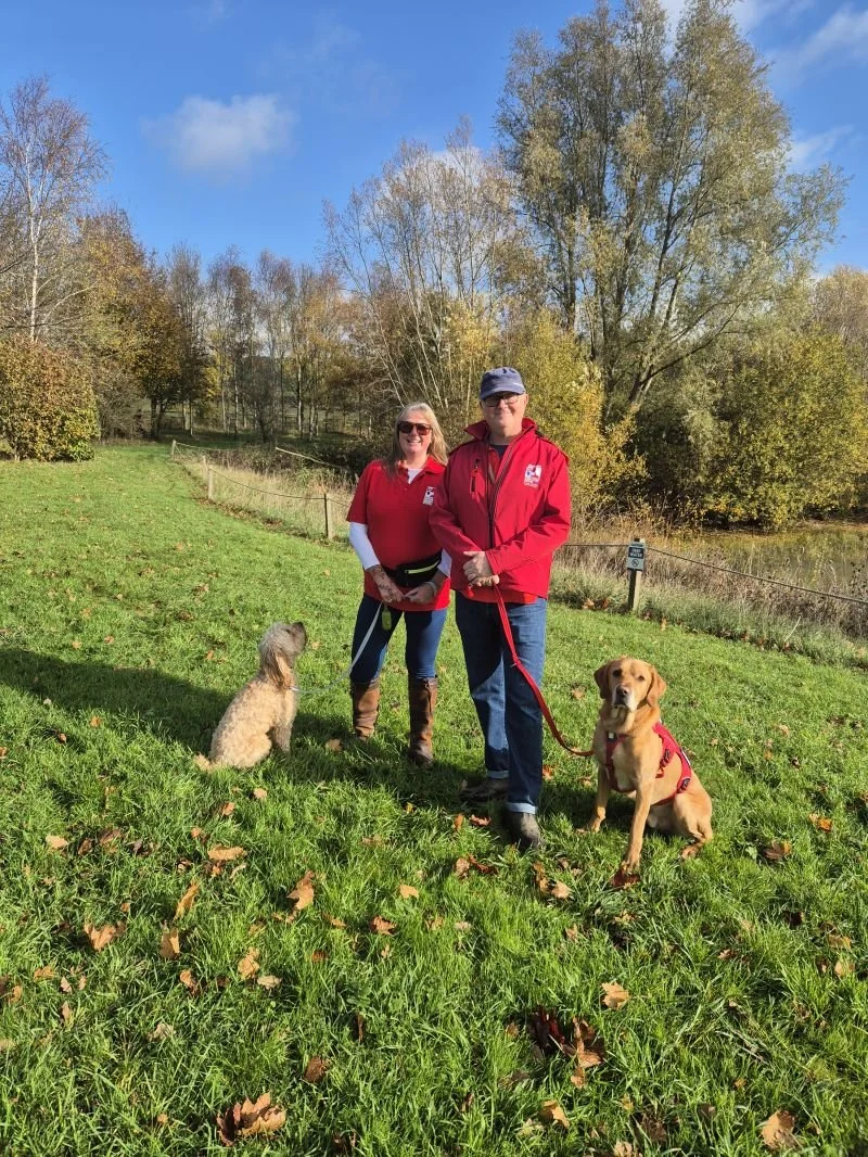 A woman and a man wearing red jackets and sunglasses standing on a grassy field with two dogs, one sitting and one lying down, near a pond and trees with fall foliage under a blue sky.