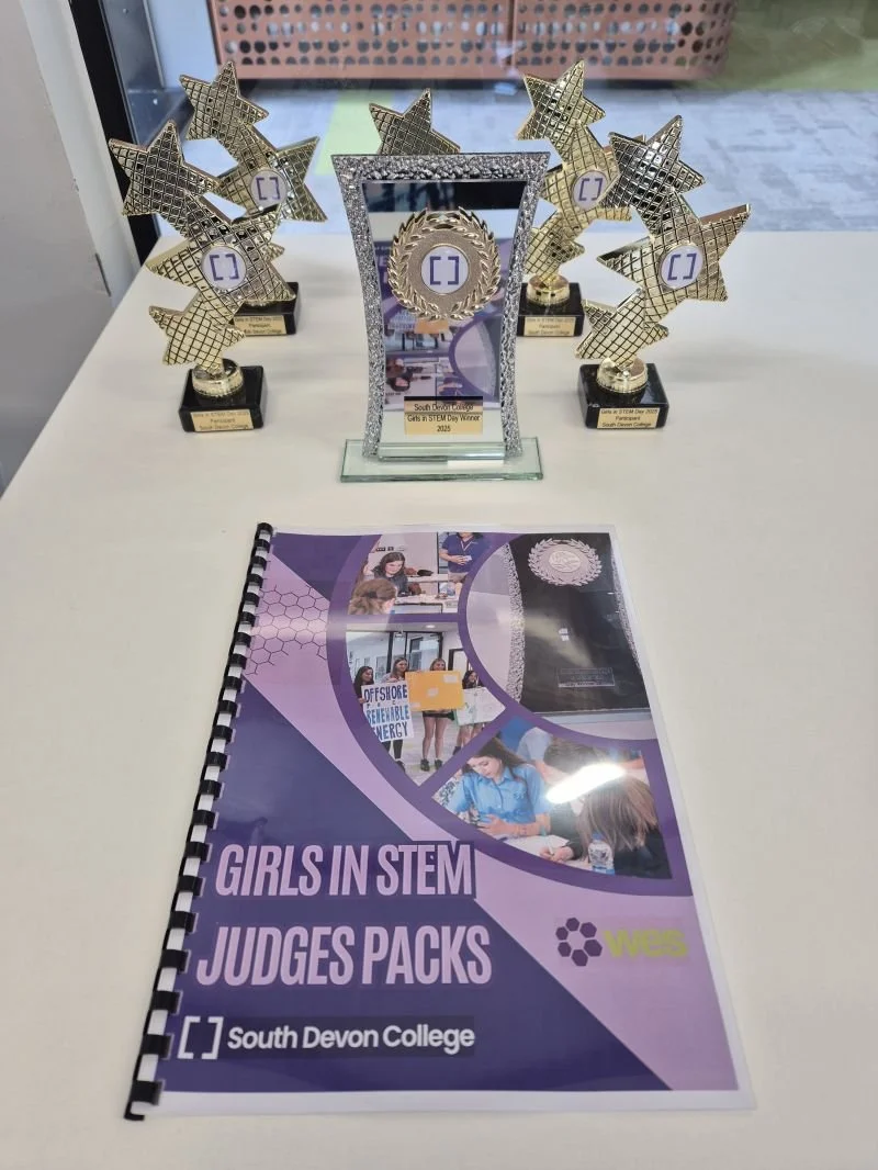 Trophies and awards arranged on a table, including a central silver frame trophy surrounded by star-shaped trophies, with a booklet titled ‘Girls in STEM Judges Packs’ from South Devon College placed in front.