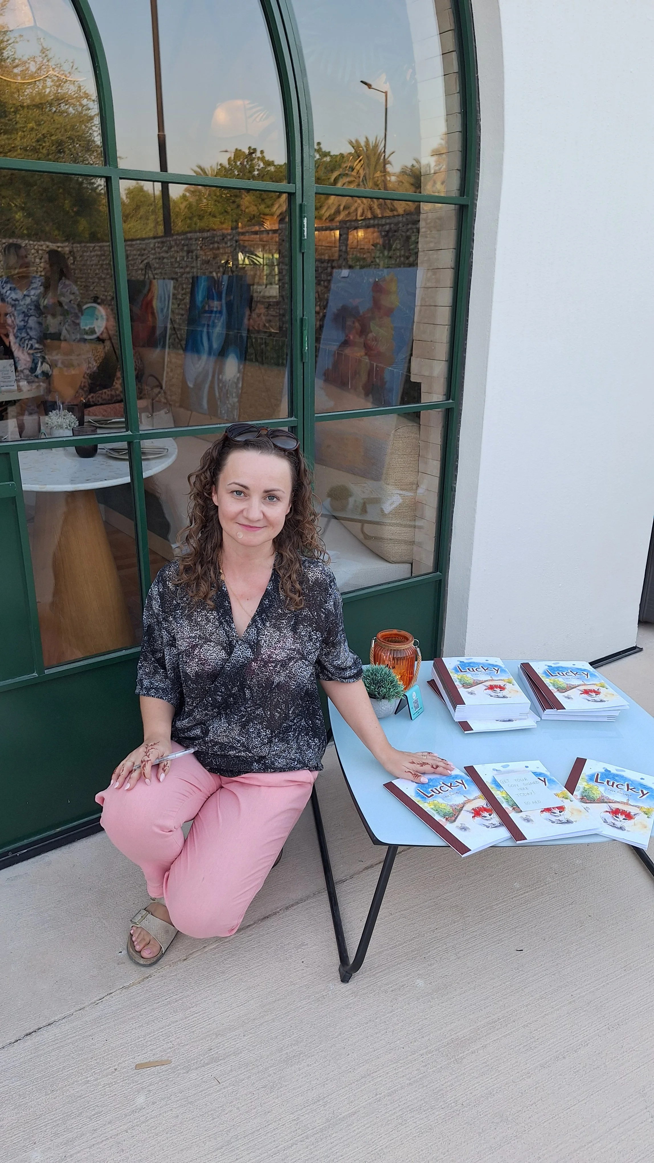 Woman sitting at a table outdoors with pamphlets titled 'Lucky', a small plant, and a candle holder. Behind her is a glass building with reflections of trees and a painting inside.