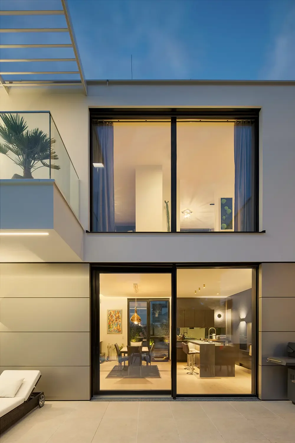 Modernes, beleuchtetes Haus mit großen Glasfenstern, Blick in ein helles Wohnzimmer und Esszimmer, Außenbereich mit Liegestuhl