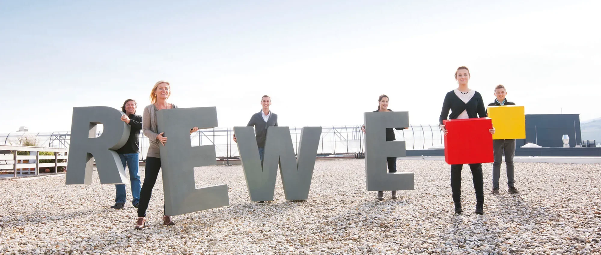 Sieben Menschen halten die Buchstaben des Wortes 'REWE' in unterschiedlichen Farben und Größen auf einem Dach, mit einem blauen Himmel im Hintergrund.