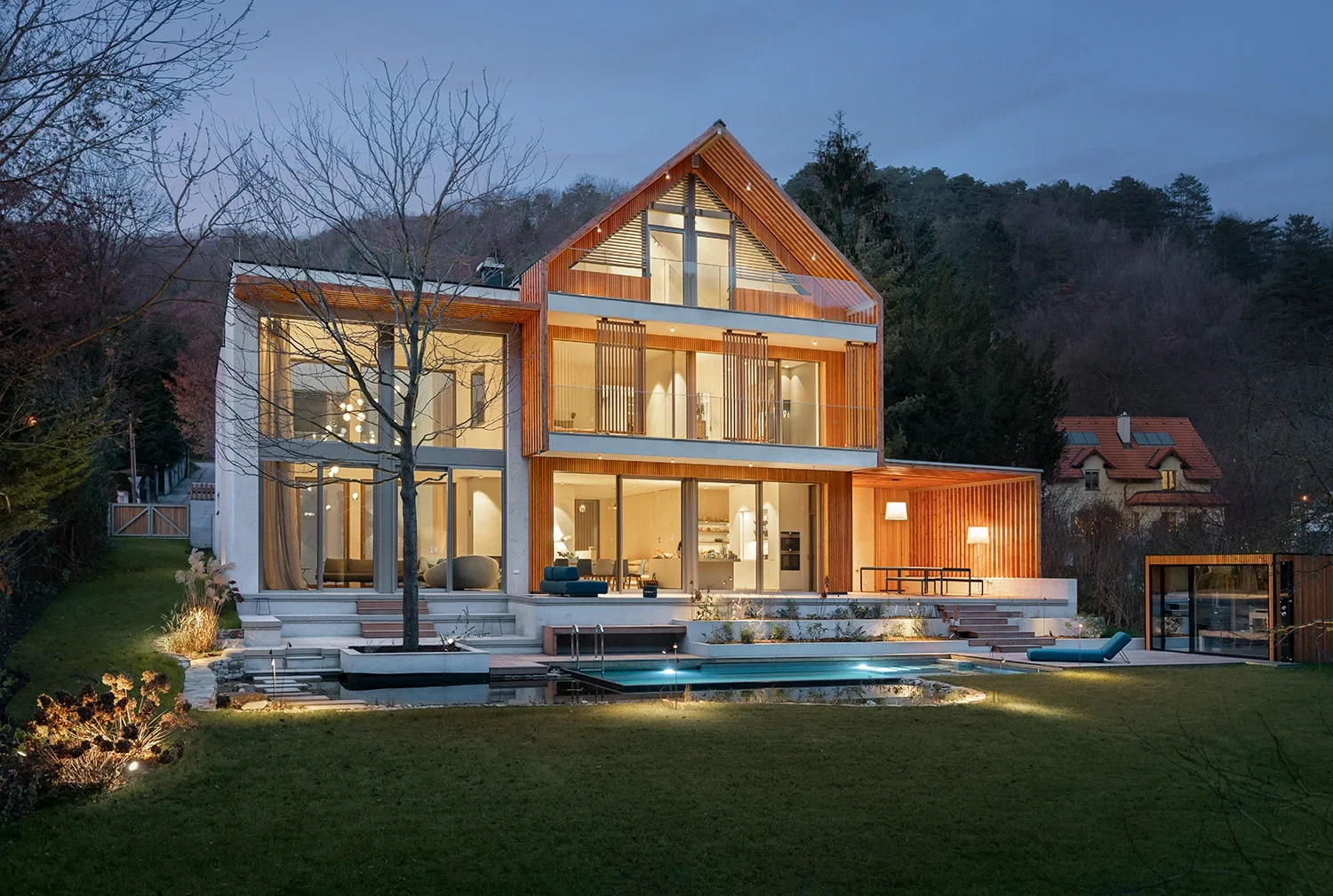Ein modernes, mehrstöckiges Haus mit großen Fenstern, Holzverkleidung und einem Pool im Garten bei Dämmerung.