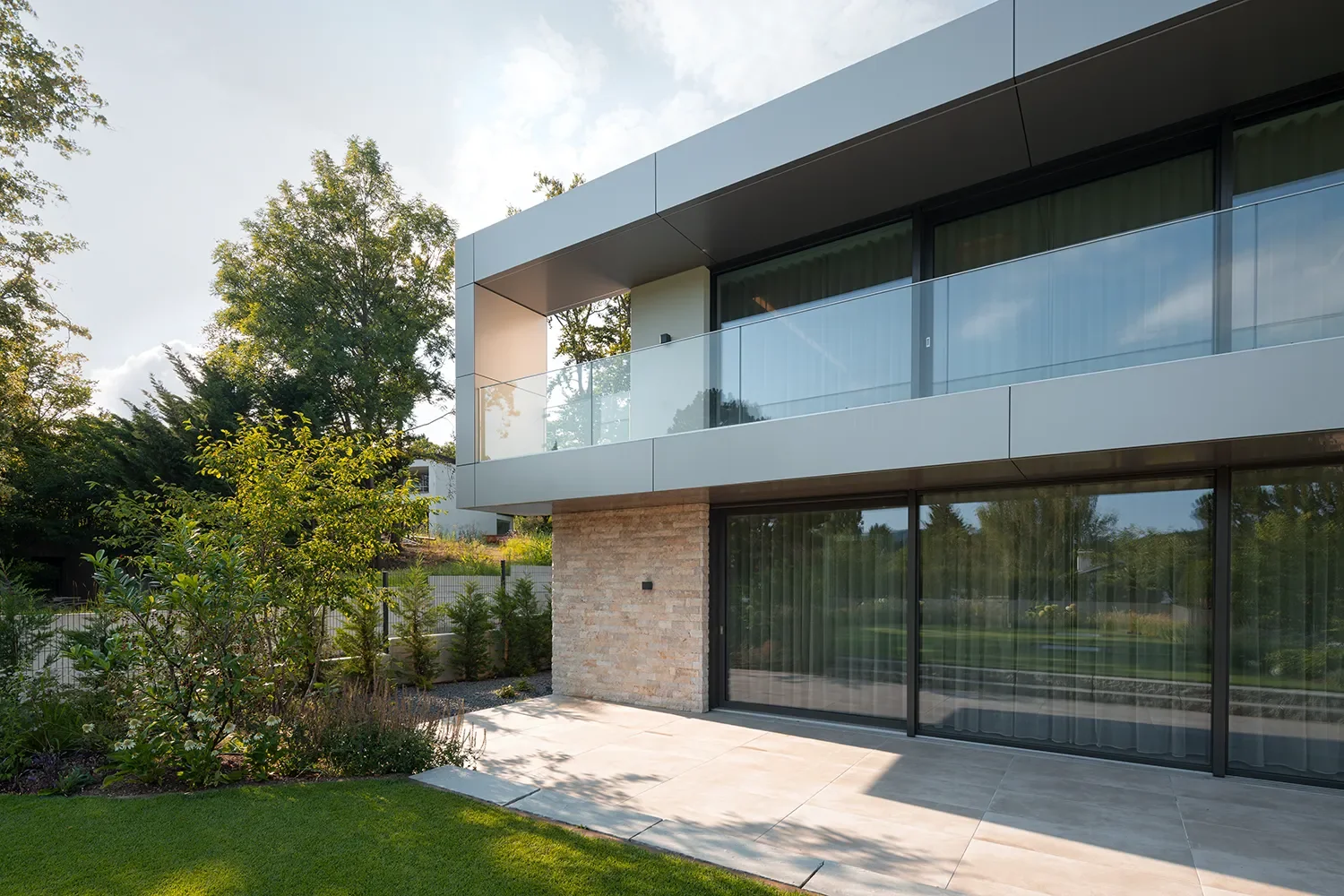 Moderne zweistöckige Hausfassade mit Glasbalustrade, große Glasfenster und Garten mit Bäumen bei Tageslicht.