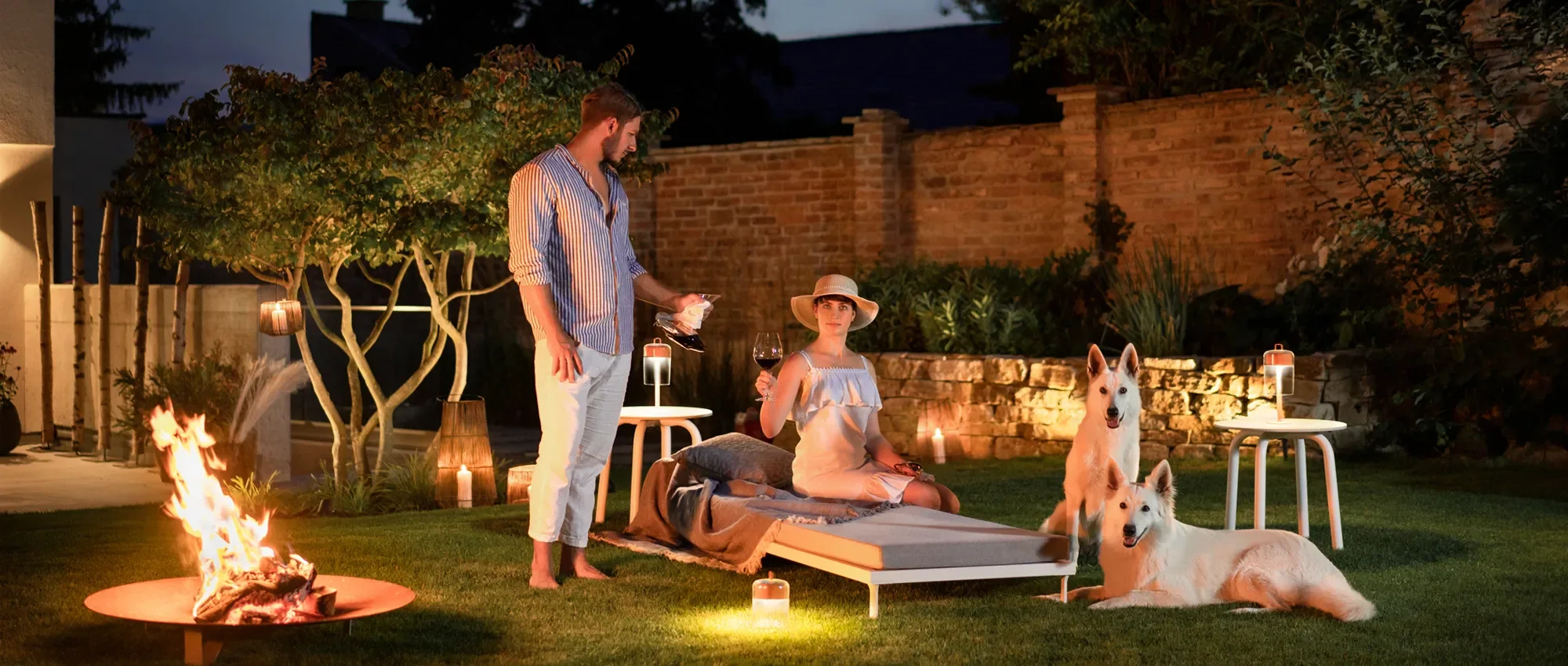 Ein Mann und eine Frau genießen einen Abend im Garten mit einem Lagerfeuer, Wein und Kerzen, begleitet von zwei weißen Hunden.