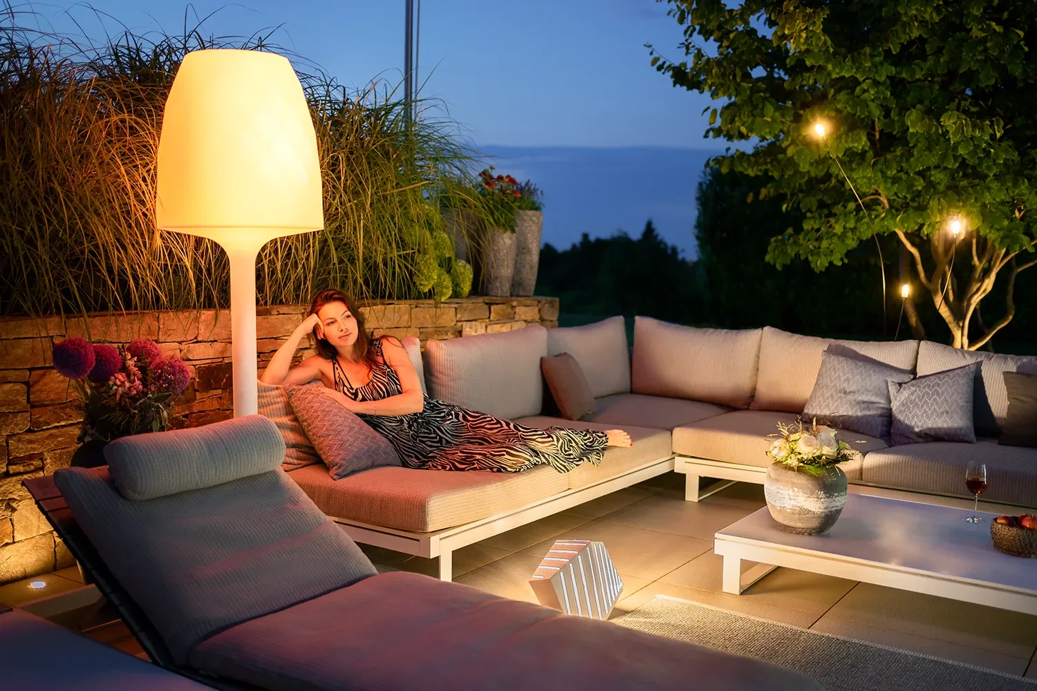 Eine junge Frau liegt entspannt auf einem gemütlichen Außen-Couchbereich, umgeben von Kissen und Lampen, bei Sonnenuntergang oder Nacht, mit Blick auf einen Himmel und Bäume im Hintergrund.