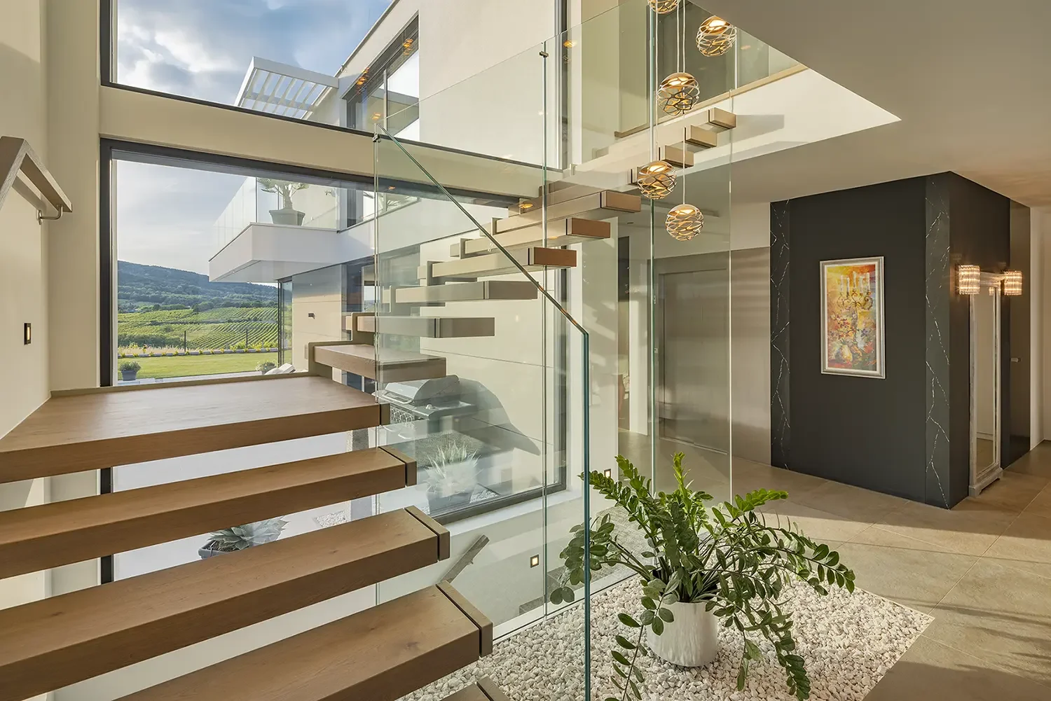 Moderner Wohnbereich mit einer Treppe aus Holz und Glas, großer Fensterfront mit Blick auf Weinberge, dekorative Beleuchtung und ein Kunstbild an der Wand.