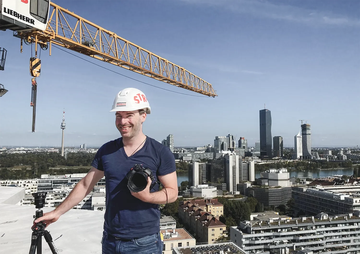 Ein Mann trägt einen weißen Bauhelm mit dem Schriftzug 'STRABAG' und hält eine Kamera sowie ein Stativ auf einem Hochhaus in Berlin, mit der Berliner Skyline im Hintergrund, einschließlich Fernsehturm und verschiedenen Wolkenkratzern bei sonnigem Himmel.