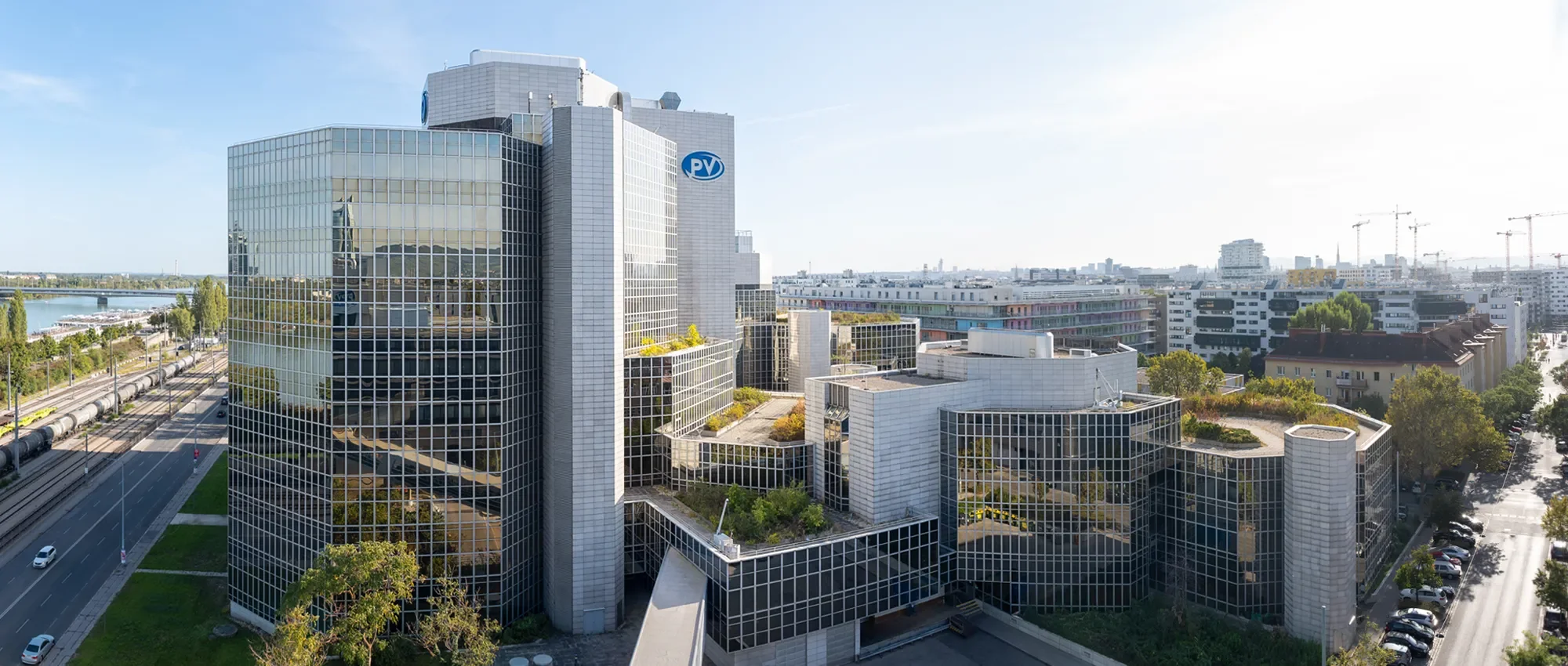 Moderne, glasverkleidete Bürofassade eines Hochhauses mit PV-Logo, umgeben von anderen Gebäuden, Straßen, Bäumen und einem Fluss im Hintergrund. 