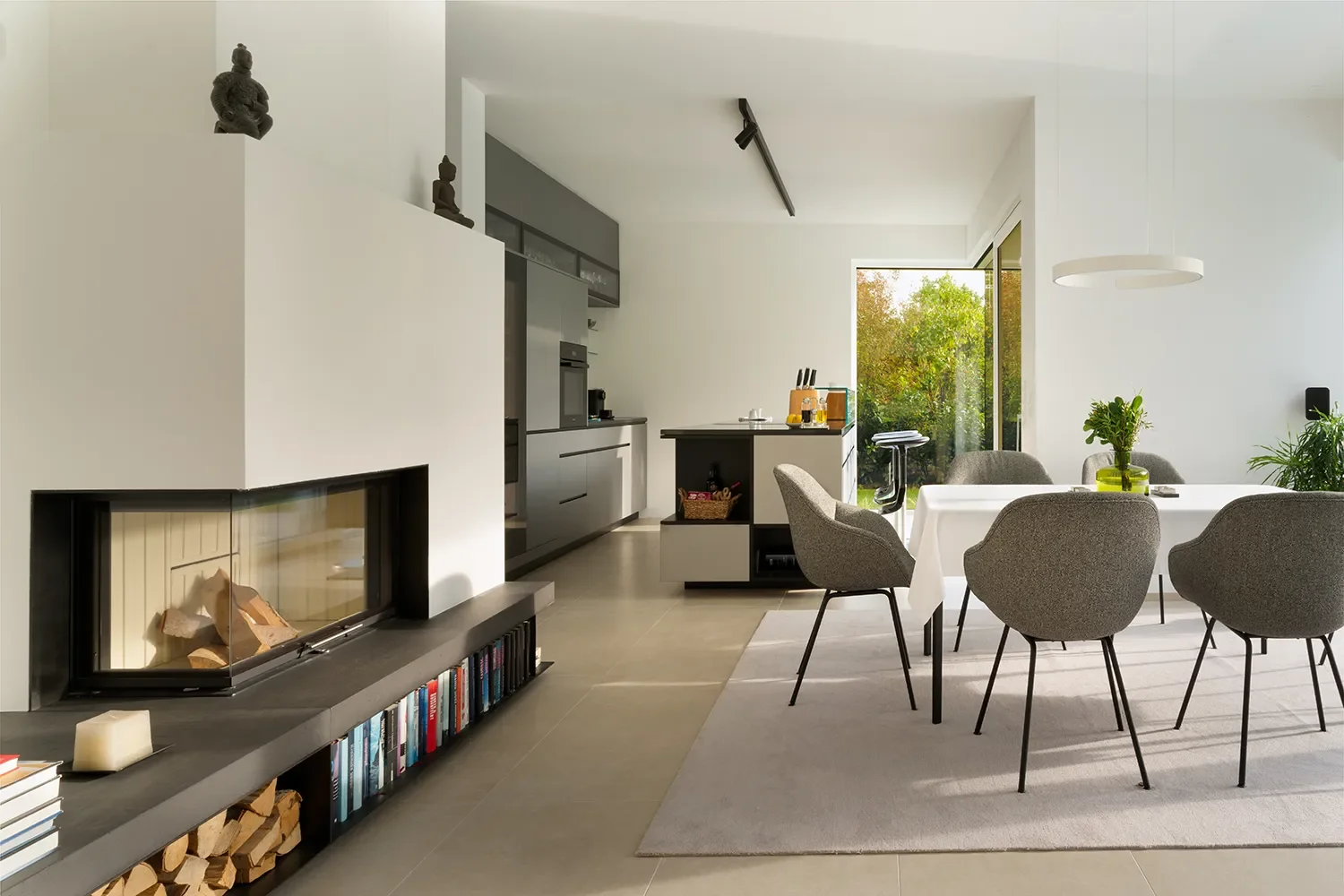 Moderne offene Wohnküche mit Essbereich, Kamin, Bücherregal mit Holzscheiten, Küchentheke mit Küchenutensilien, große Fenster mit Blick auf Garten, Pflanzen und natürliche Beleuchtung.