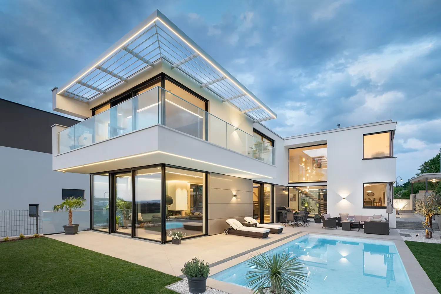 Moderne, weiße Villa mit großer Terrasse, Glasgeländer, Swimmingpool und Garten bei Abenddämmerung