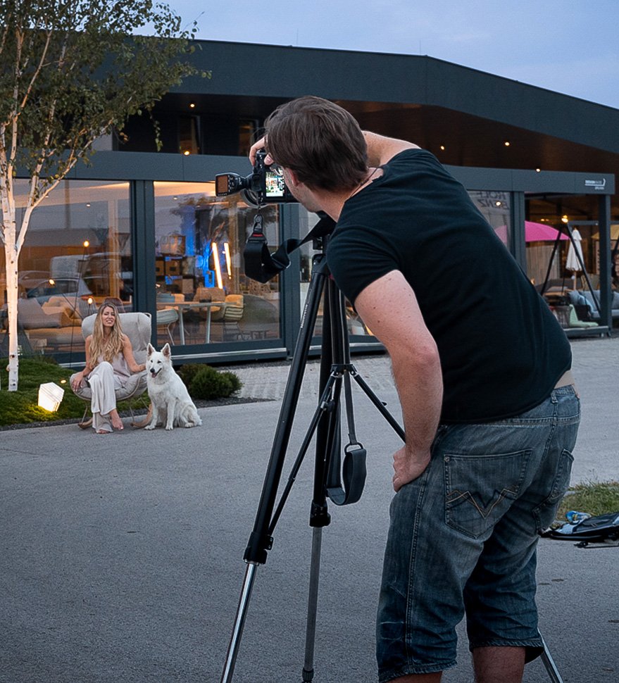 Ein Fotograf macht ein Fotoshooting im Freien bei Dämmerlicht. Eine junge Frau sitzt auf einem Stuhl mit einem weißen Hund neben ihr, vor einem modernen Gebäude im Hintergrund.